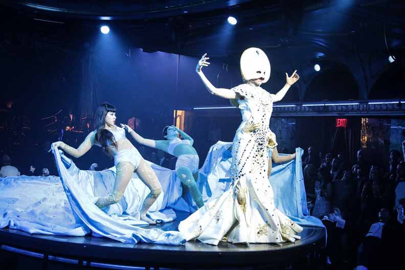 Queen of the Night, Dancer  Katherine Crockett  wearing the mask which is round half mask, that is large with a jewel at top of the mask.   Wearing a long jewel dress with 3 dancers holding the ends of the dress.  